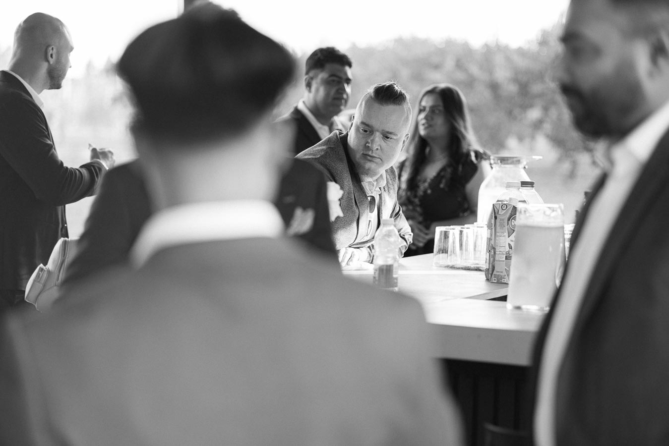 Black and white photo of people socializing at a bar, focusing on a man in a suit leaning on the counter with other people in the background.