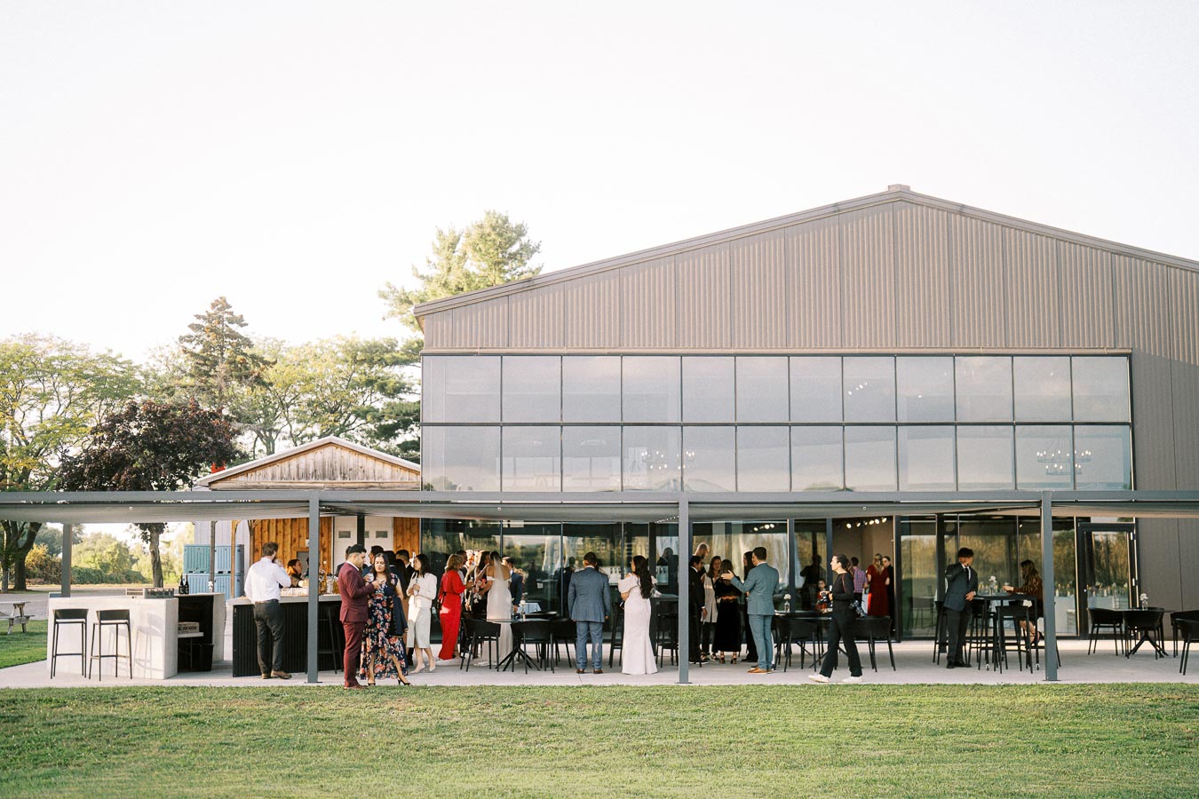 Outdoor event at a modern venue with glass windows, featuring guests mingling in formal attire under a covered patio area.