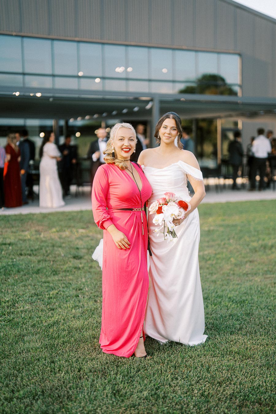 Two women posing outdoors at an event, one wearing a pink dress and the other in a white wedding gown holding a bouquet, with a modern building and attendees in the background.