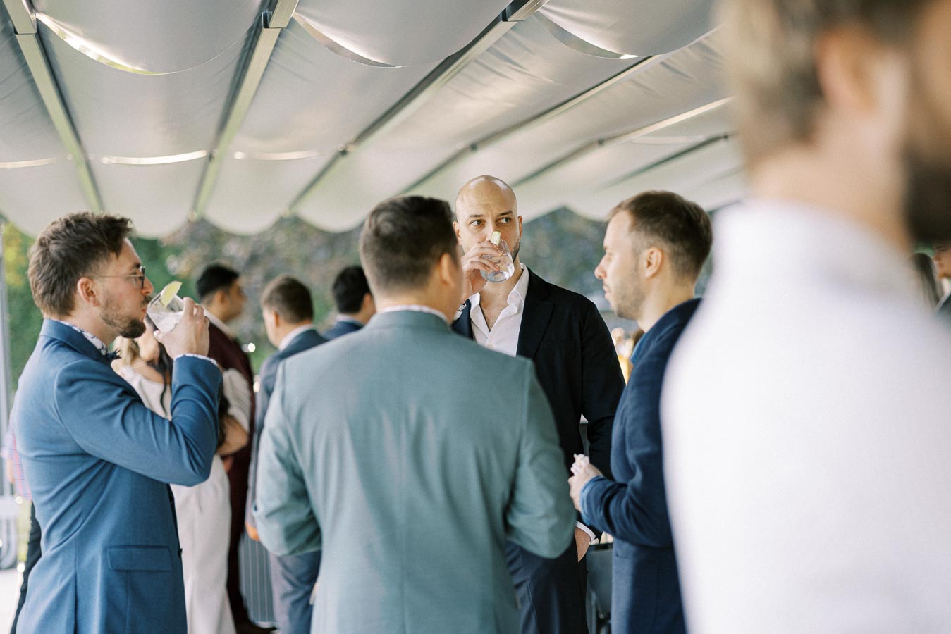 A group of elegantly dressed men socializing at an outdoor event, with one man prominently sipping a drink while engaged in conversation under a modern canopy.