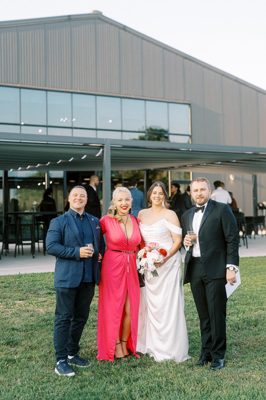 A woman in a wedding dress stands with three people in formal attire outside a modern building. The man in casual attire holds a drink, the woman in a pink gown smiles, and the man in a tuxedo has a drink as well, all posing on green grass.