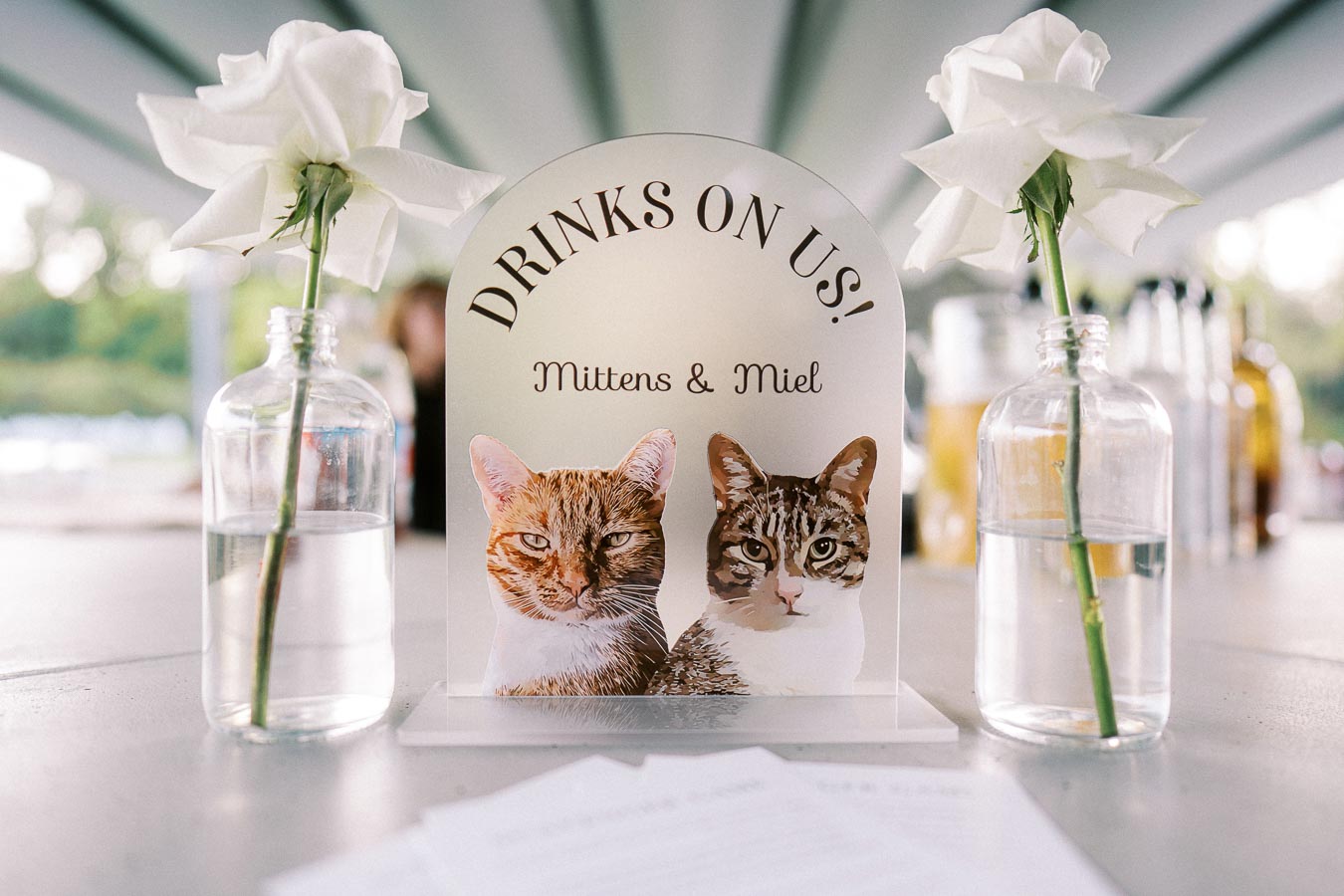 A personalized wedding sign featuring two cats with the text Drinks On Us! Mittens & Miel, placed between two glass vases with white roses, set on a table at an outdoor event.