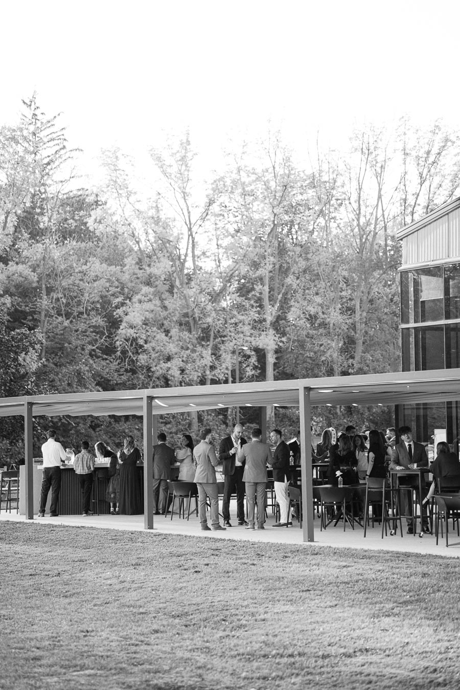 Black and white image of a group of people socializing at an outdoor event under a modern patio structure, with trees in the background. Some are seated at tables, while others stand, dressed in formal attire.
