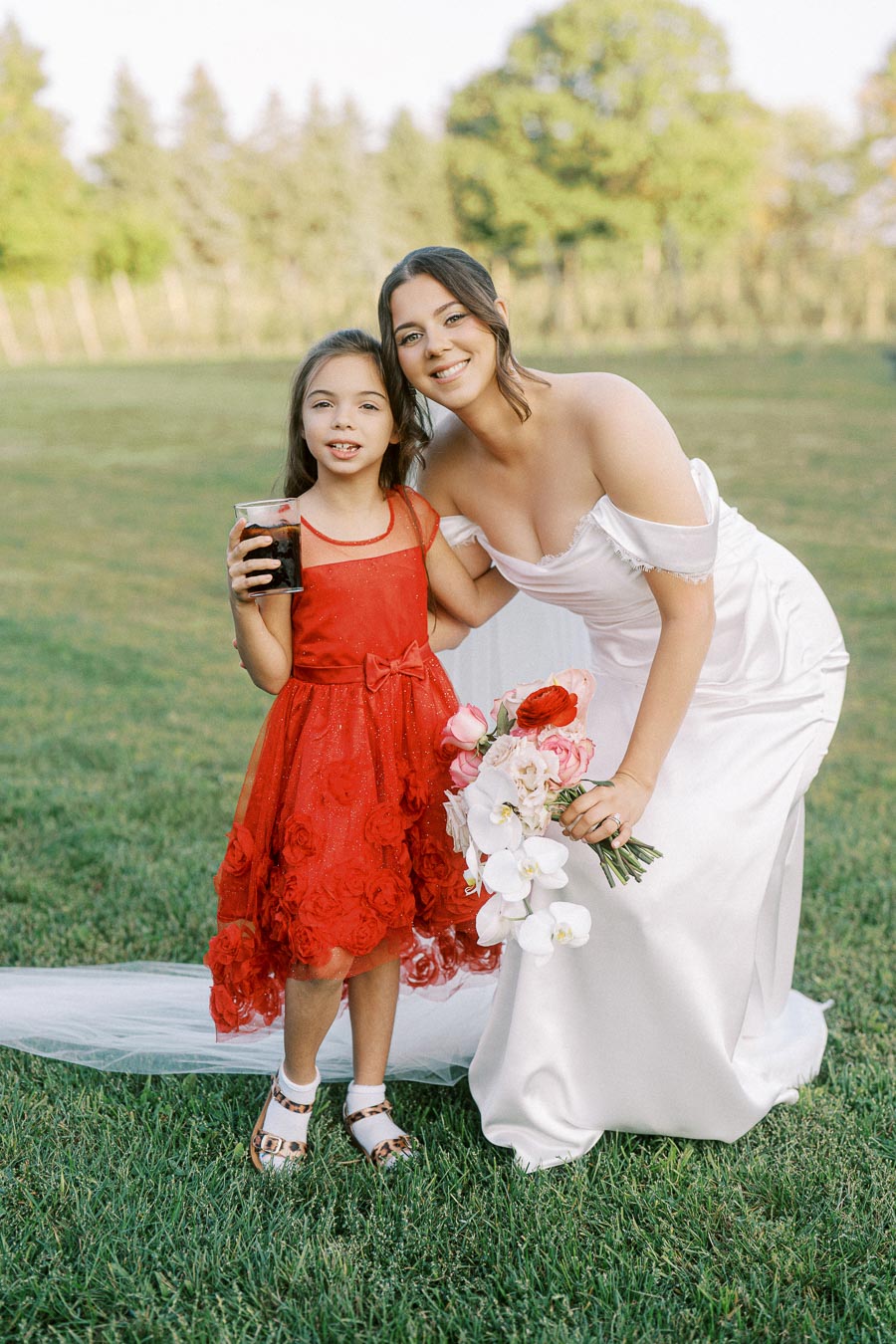 A bride in a white wedding dress smiles while posing with a young girl in a red dress holding a drink, standing together on green grass with trees in the background. The bride holds a bouquet of pink, white, and red flowers.