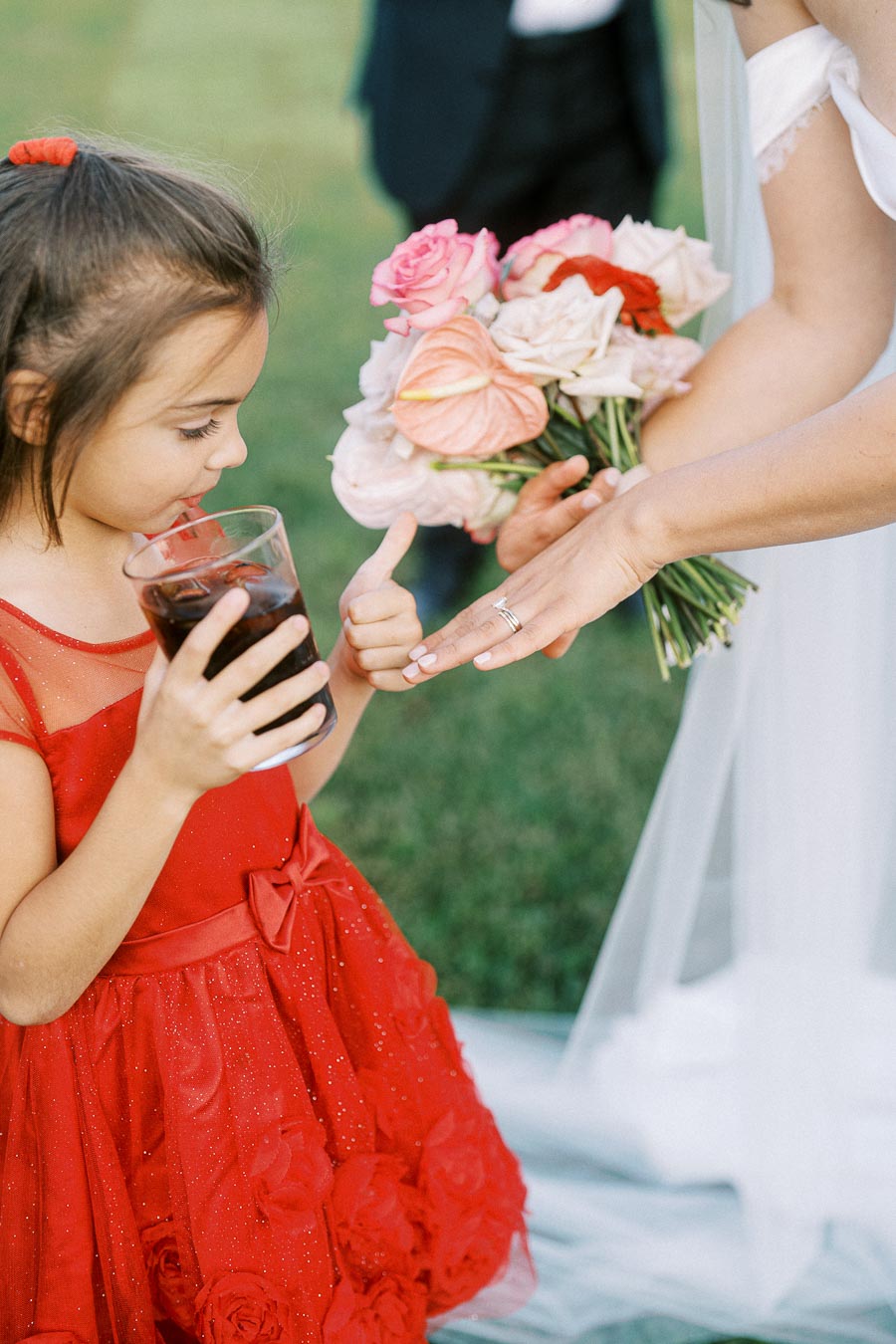A young girl in a red dress holding a glass of soda looks at a bride's hand and wedding ring, with the bride holding a bouquet of colorful flowers in an outdoor setting.