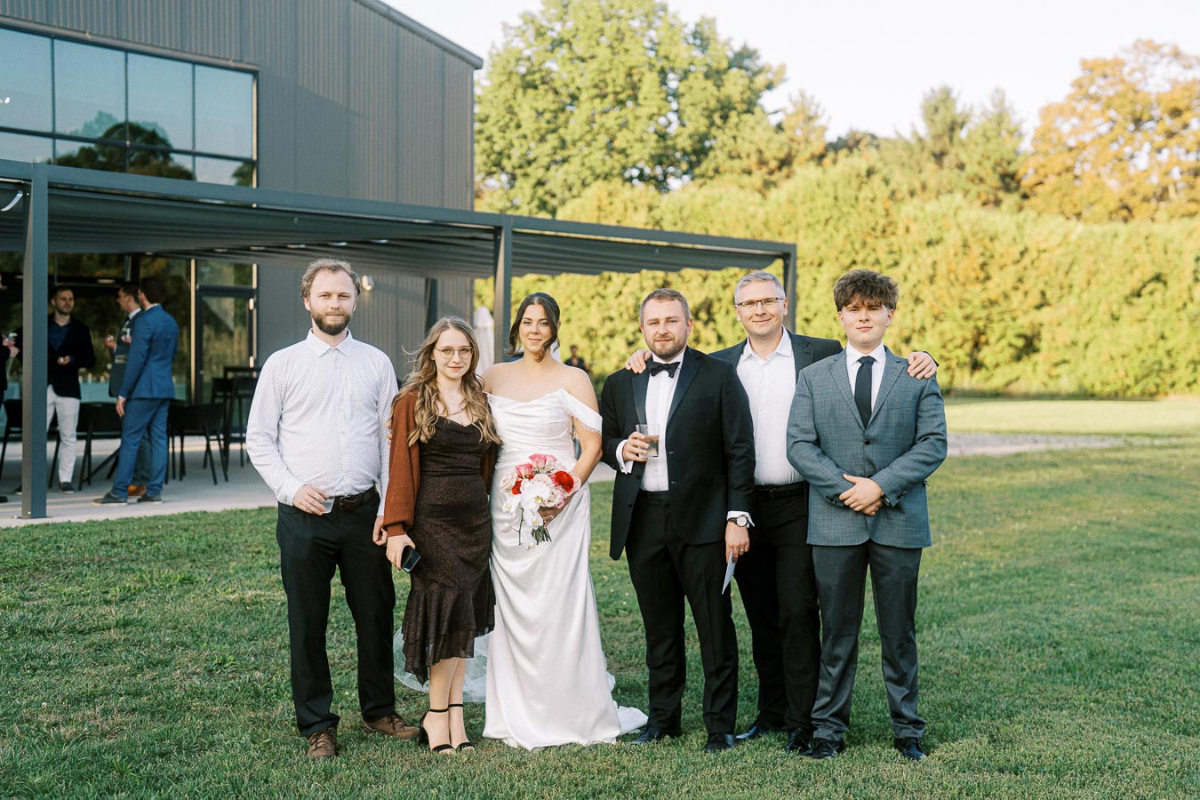 Group photo of a wedding party standing outdoors in formal attire, featuring a bride in a white dress holding a bouquet, with friends and family in suits and dresses, in front of a modern building and greenery.