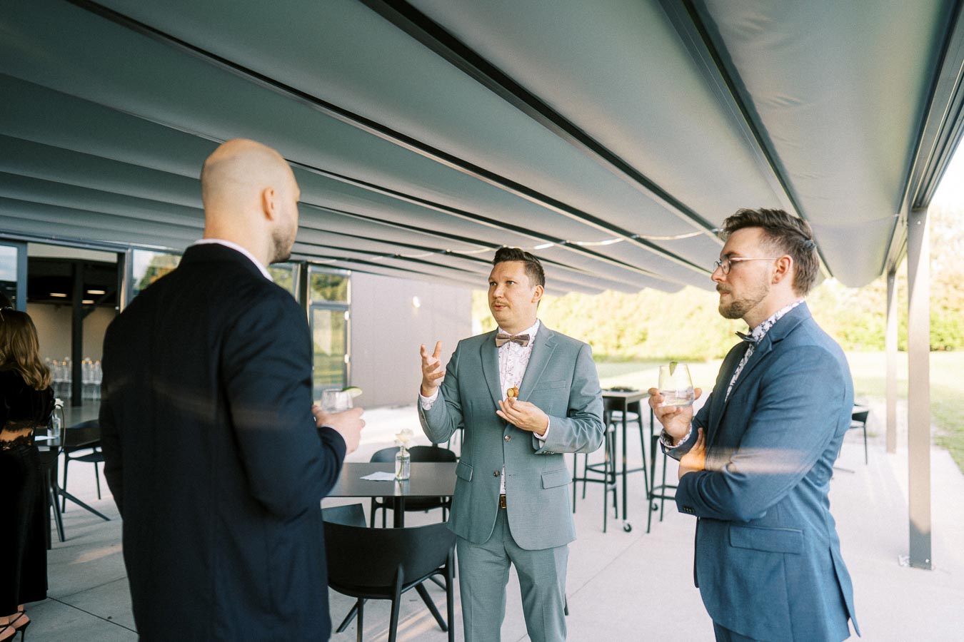 Three men in formal suits engaging in conversation during an outdoor event under a large canopy, holding drinks and appetizers.