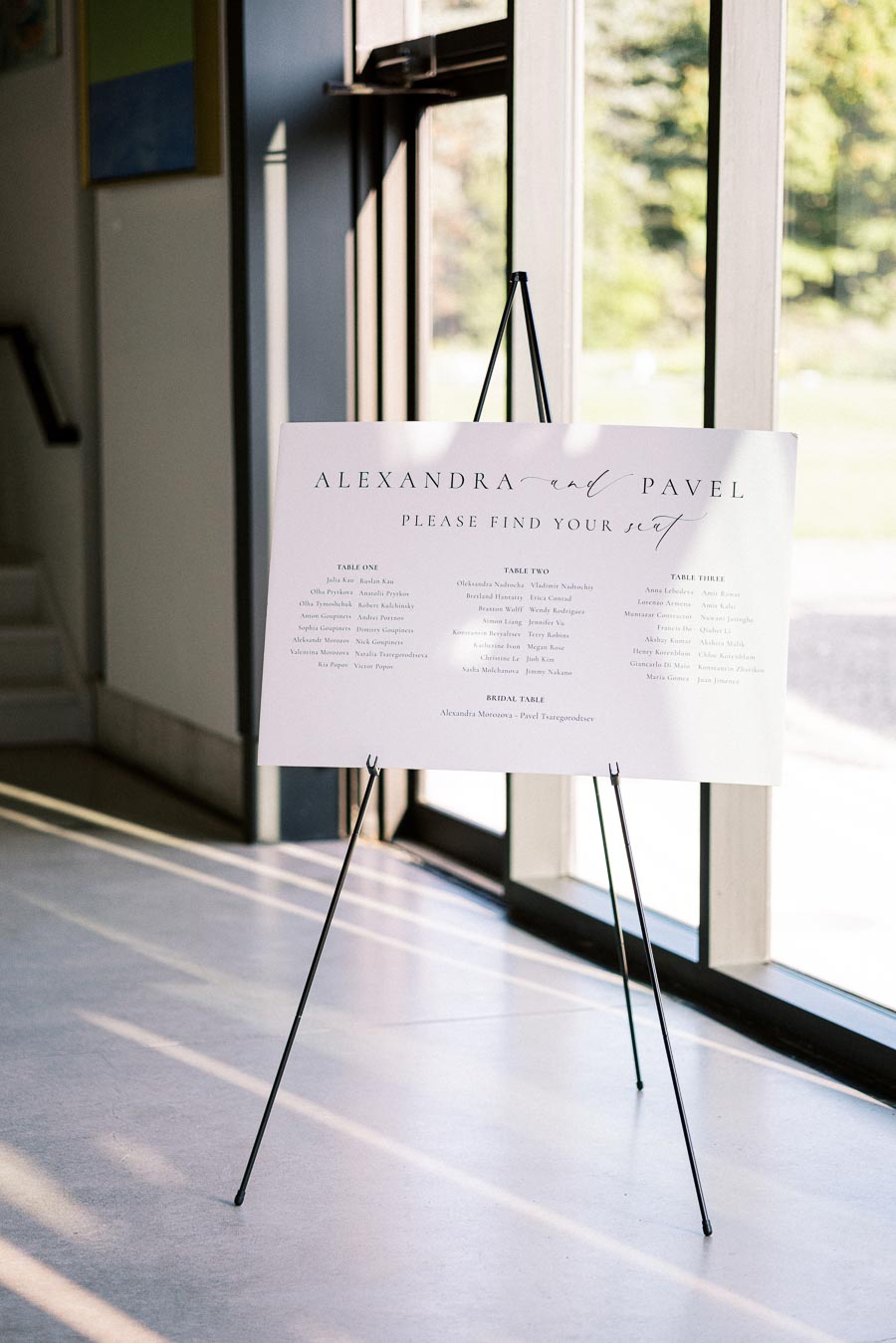 Wedding seating chart on an easel by a window, displaying guest arrangements for Alexandra and Pavel's event.