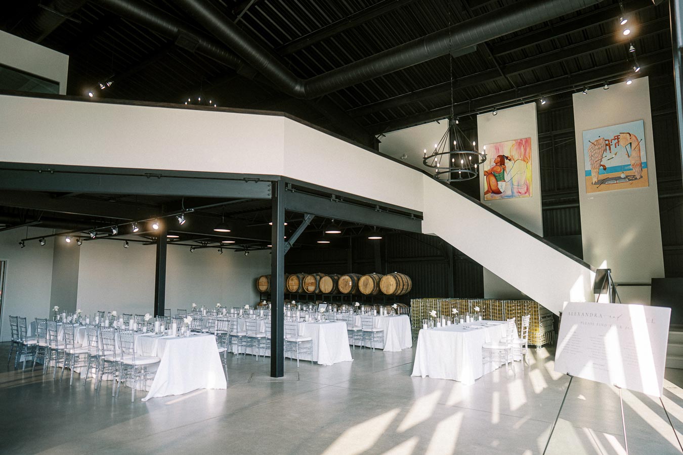 Modern industrial event space with elegant white table settings and barrels, featuring high ceilings, contemporary lighting, and large artworks on the walls, perfect for weddings or corporate gatherings.
