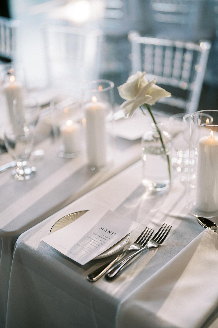 Elegant wedding table setting with white candles, a single white rose in a glass vase, menu card, and silver cutlery on a white tablecloth.