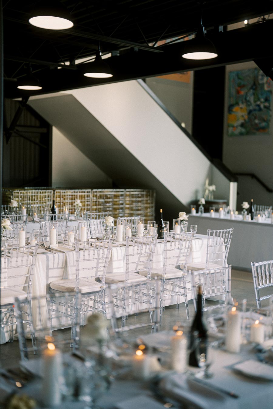 Elegant wedding reception venue with sleek, transparent chairs and tables draped in white linens, adorned with candles and wine bottles, creating a sophisticated and romantic atmosphere.