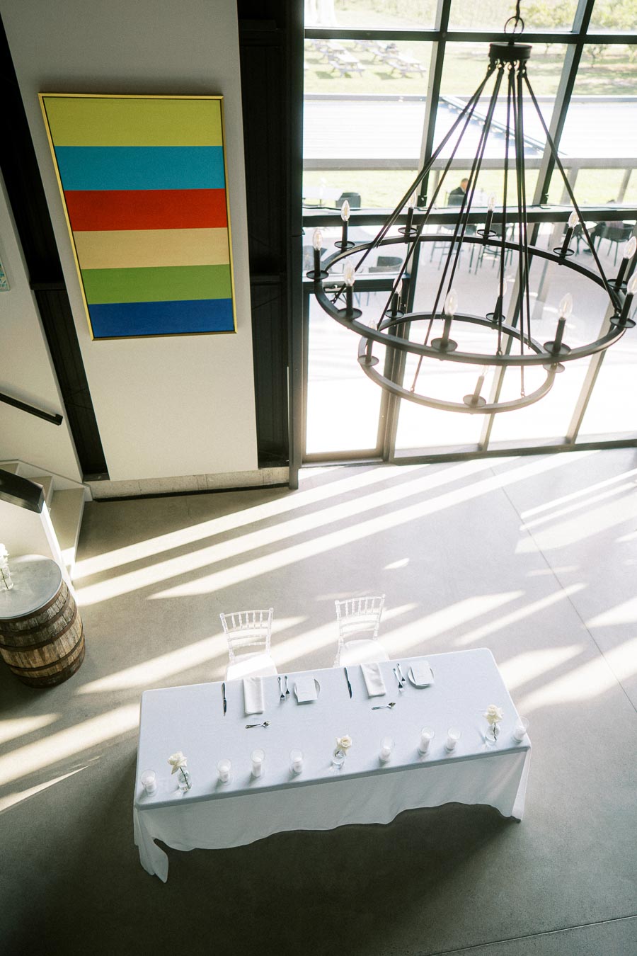Modern interior with a minimalist setup, featuring a long dining table with white tablecloth and place settings, positioned under a large window. Overhead, a rustic chandelier complements the ambient light, while a colorful abstract painting adorns the wall. Ideal venue for elegant events and gatherings.