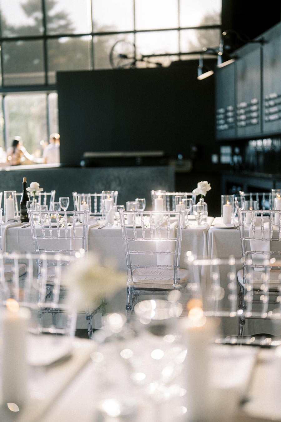 Elegant indoor wedding reception setup with white tablecloths, clear glass chairs, candles, and floral centerpieces in a softly lit modern venue.
