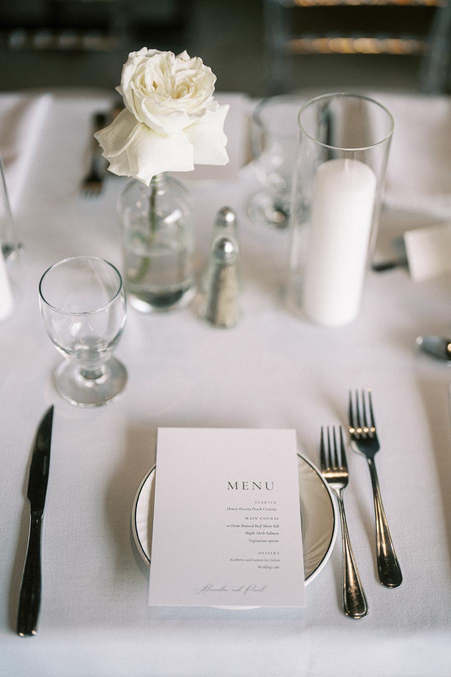 Elegant table setting with a white rose centerpiece, a menu card, silver cutlery, a glass cup, salt and pepper shakers, and a decorative white candle on a crisp white tablecloth. Perfect for a sophisticated wedding or formal dinner.