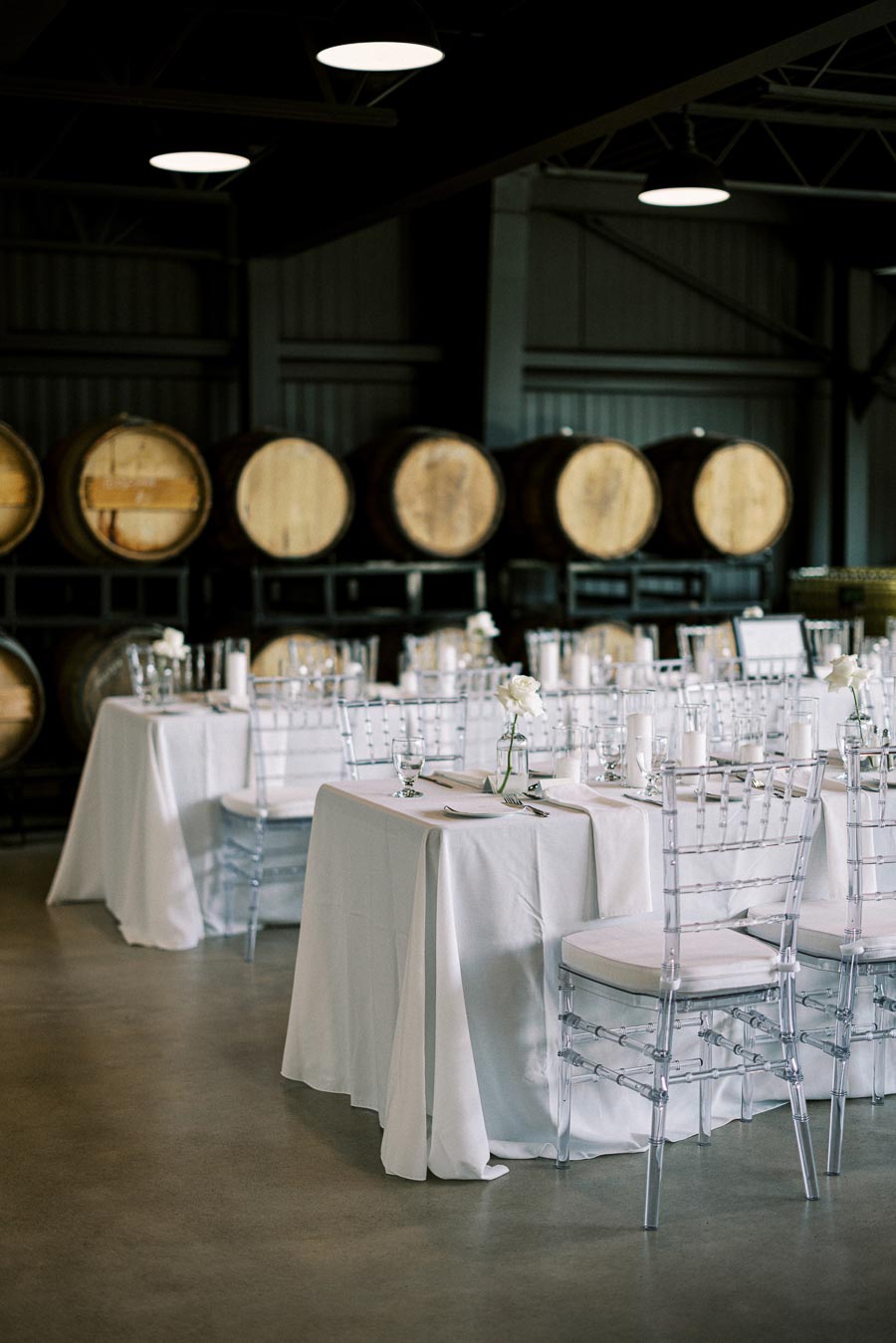 Elegant wedding reception setup in a rustic winery setting with white table linens, clear chairs, and rows of wooden wine barrels in the background.