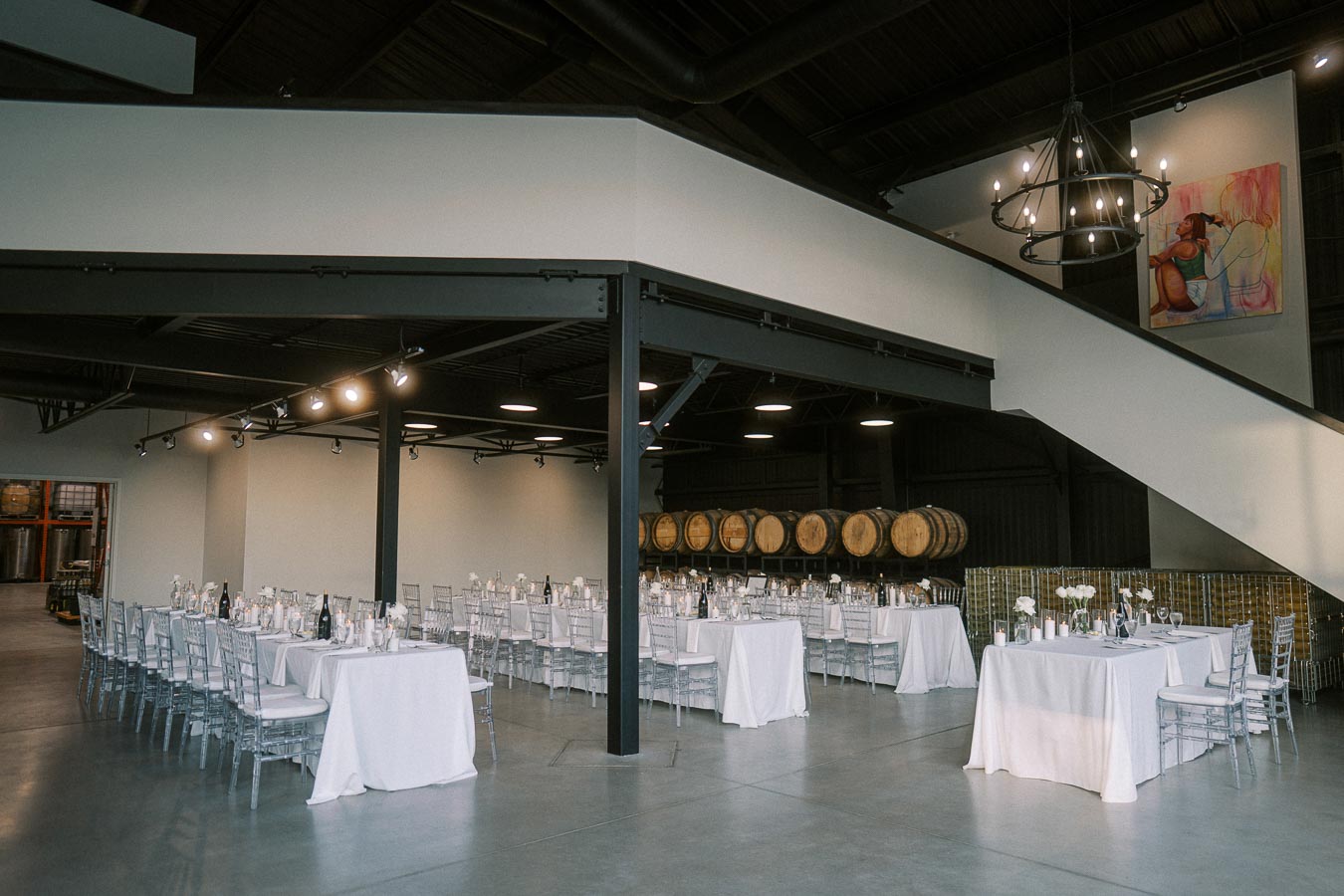 Spacious, elegantly decorated event space with white-clothed tables and silver chairs, featuring large wooden barrels and modern lighting, ideal for weddings or corporate gatherings.