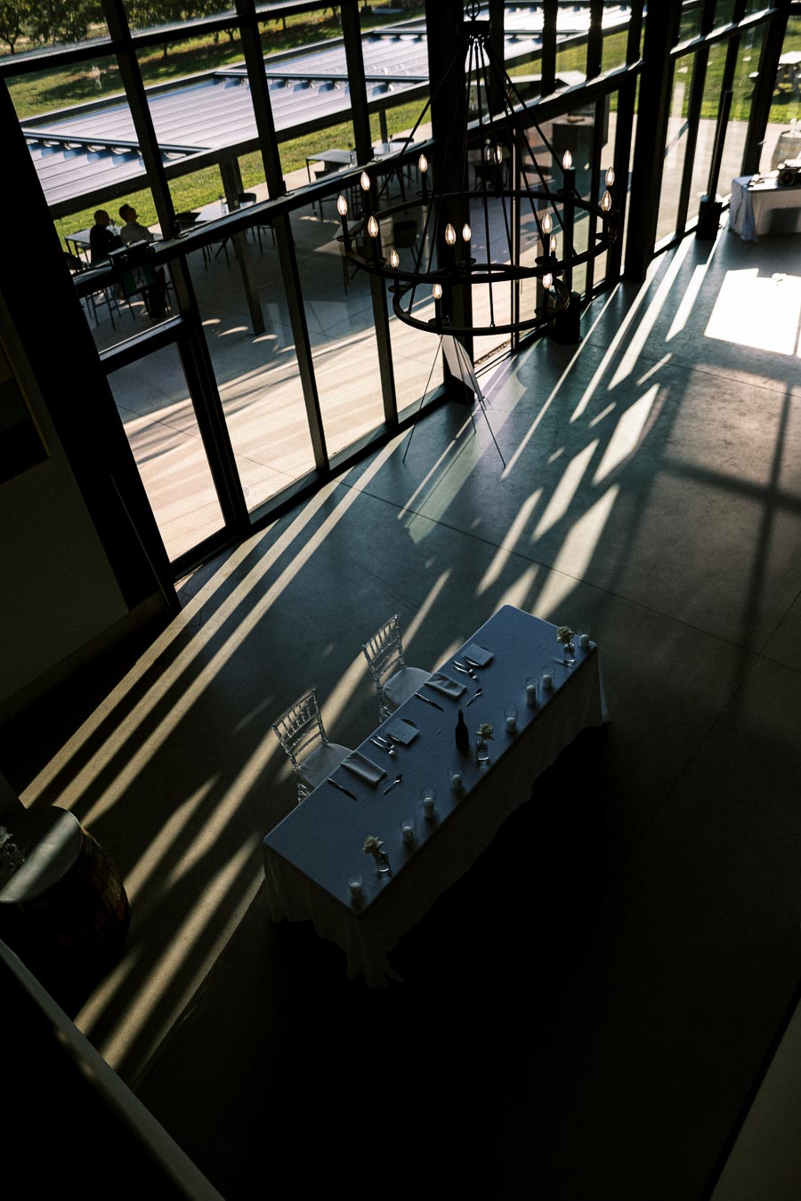 A spacious modern event venue with large glass windows allowing natural light to cast dynamic shadows. A tastefully arranged table with white linens and elegant place settings sits under a stylish chandelier.