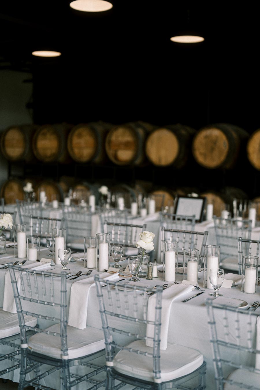 Elegant winery wedding reception setup with white table linens, clear chairs, candle centerpieces, and wine barrels in the background.