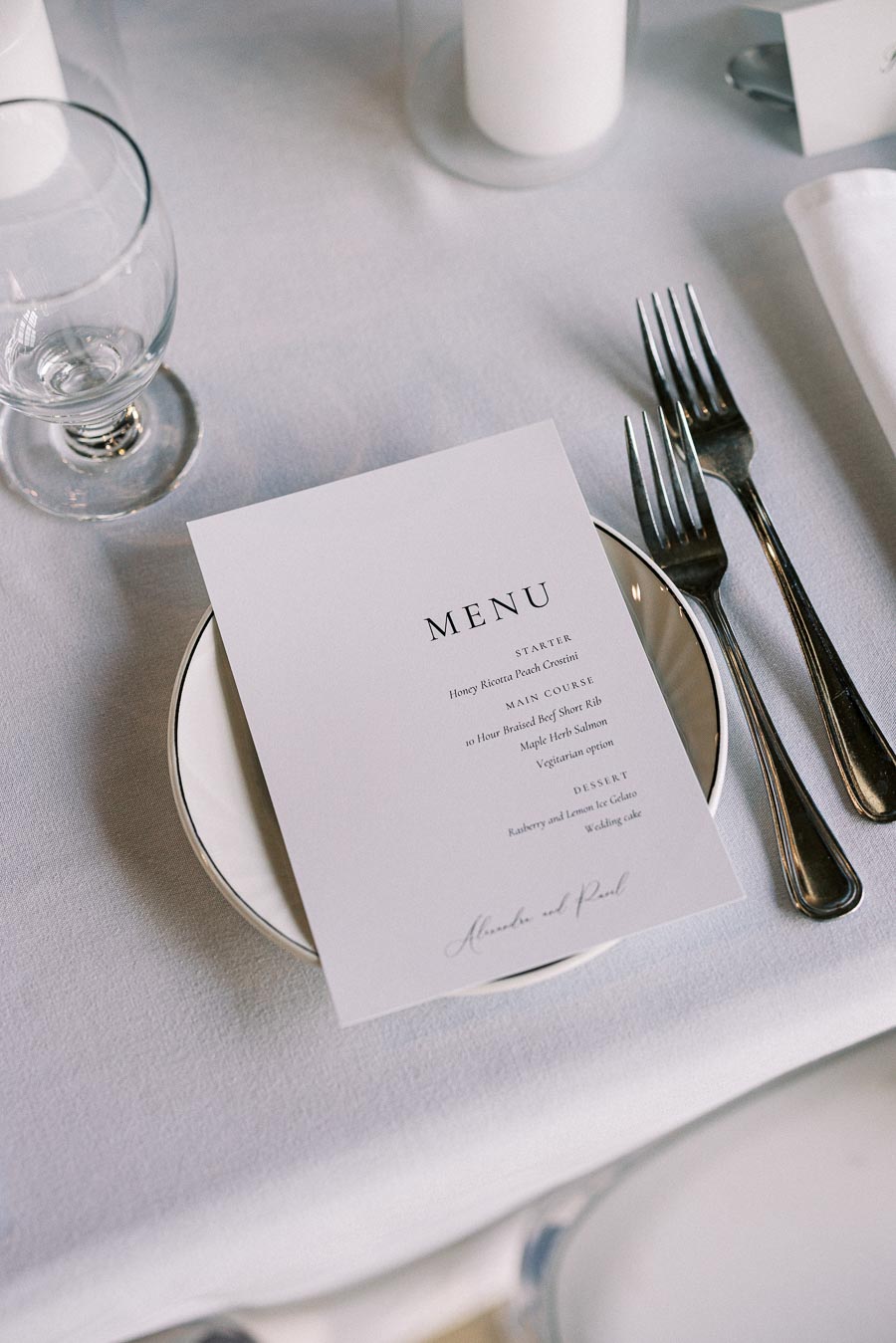 Elegant wedding table setting with a menu card displaying dinner options, placed on a white plate with polished silverware, next to a wine glass and a candle, on a white tablecloth.