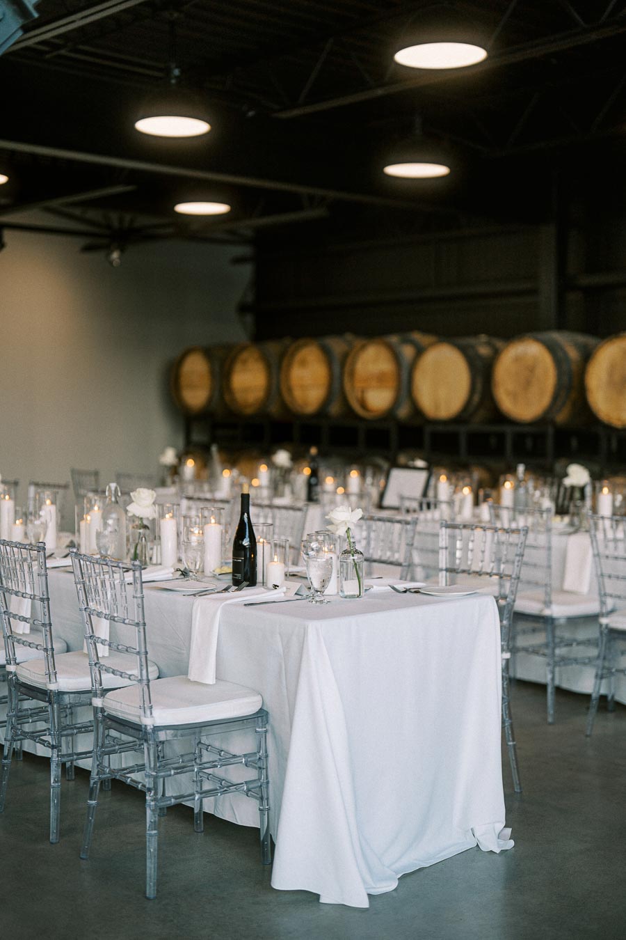 Elegant wedding reception setup in a winery with white tablecloth-covered tables, transparent chairs, candle centerpieces, wine bottle, and barrels in the background.