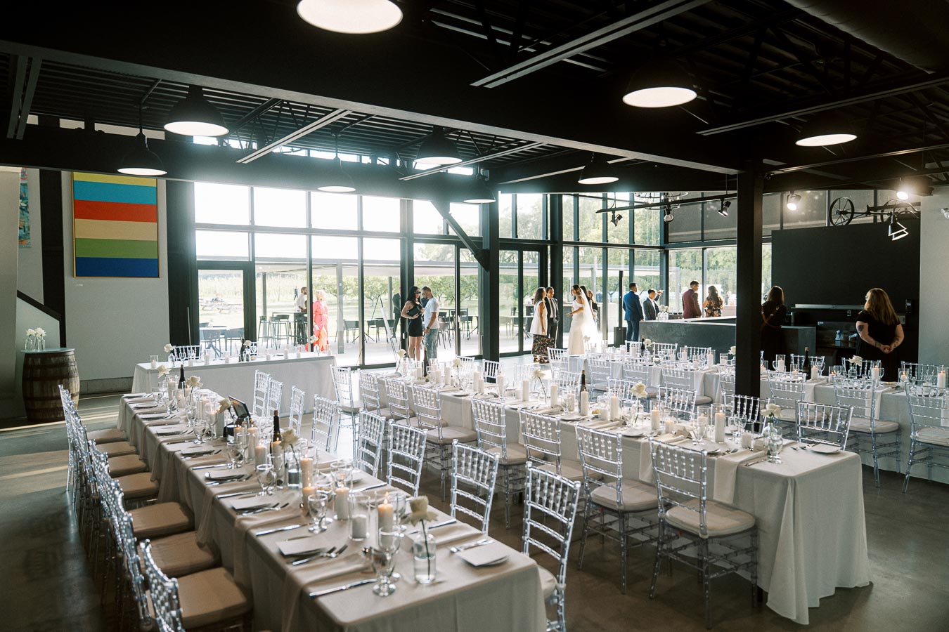 Modern wedding reception venue with elegantly set long tables and clear chairs, featuring large windows and sophisticated indoor lighting. Guests mingling in the background, creating a lively atmosphere.