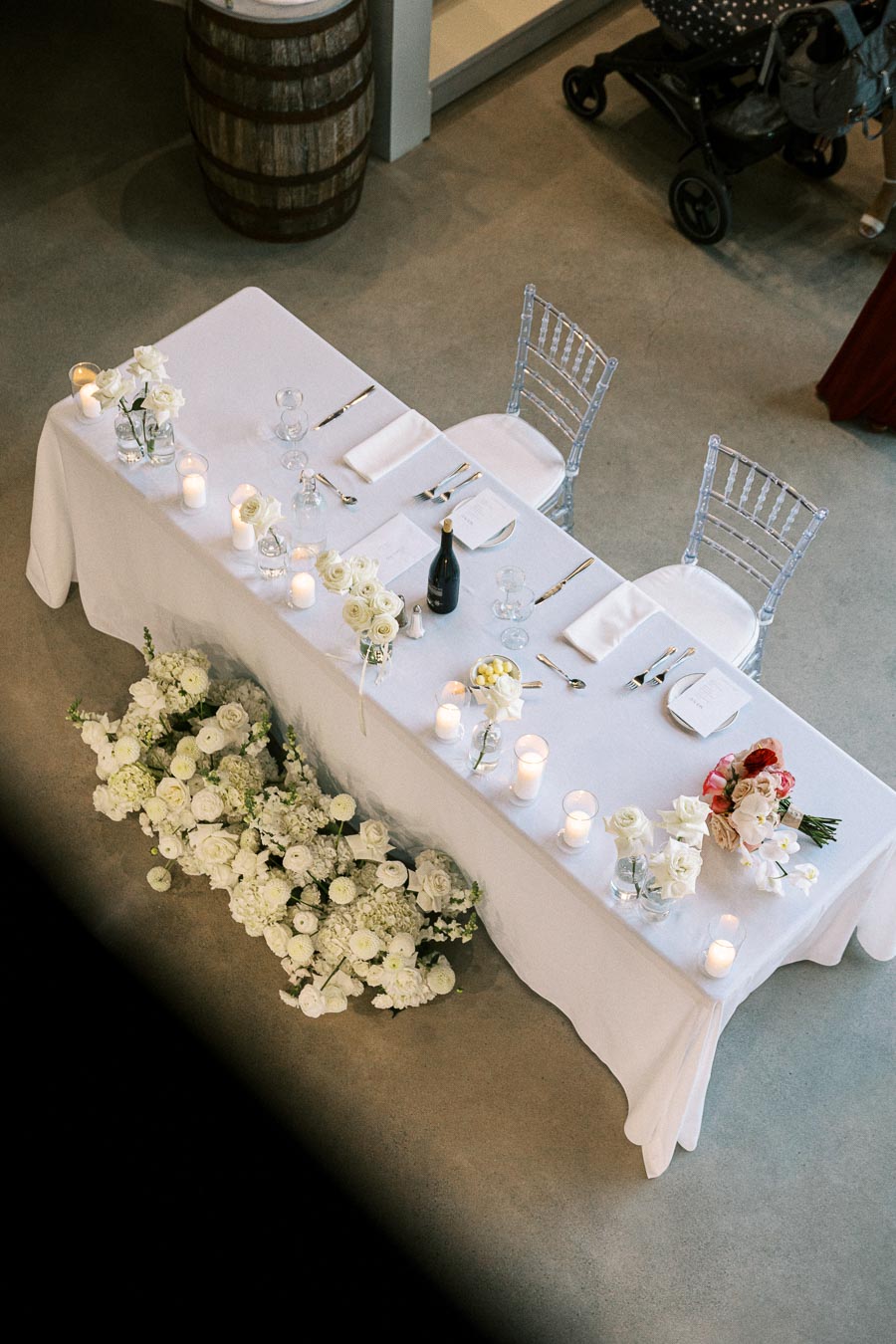 Elegant wedding table setup with white tablecloth, floral arrangements, and candlelight, viewed from above. Featuring clear chairs, cutlery, and wine bottle on the table, with a backdrop of white and pink flowers, creating a romantic ambiance.