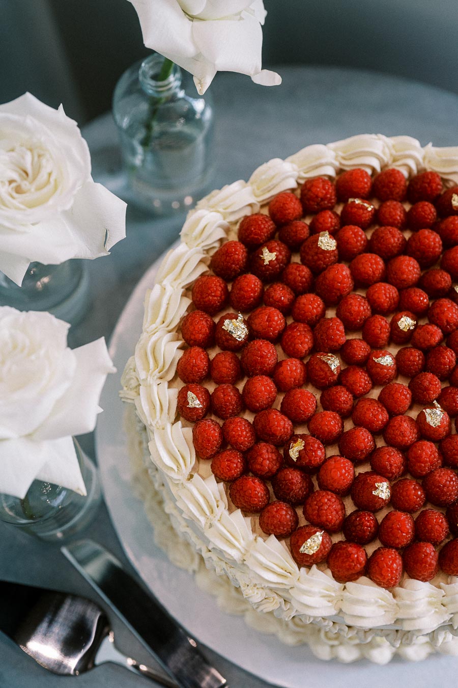 Elegant cake adorned with fresh raspberries and gold leaf, set beside white roses in glass vases, perfect for a luxurious celebration.