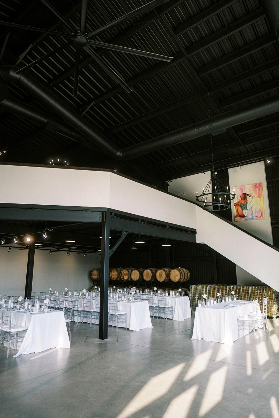 Spacious modern event venue with elegant white table settings and large industrial ceiling, featuring barrel decor and a colorful painting. Ideal for weddings, receptions, or corporate events.