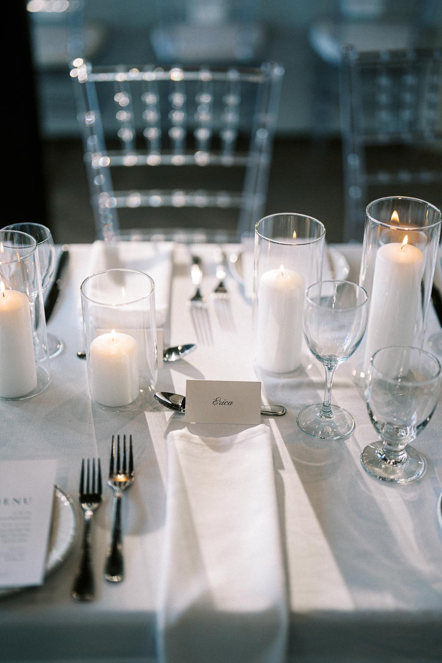 Elegant wedding table setting with white candles, glassware, cutlery, and a personalized name card Erica on a white tablecloth.