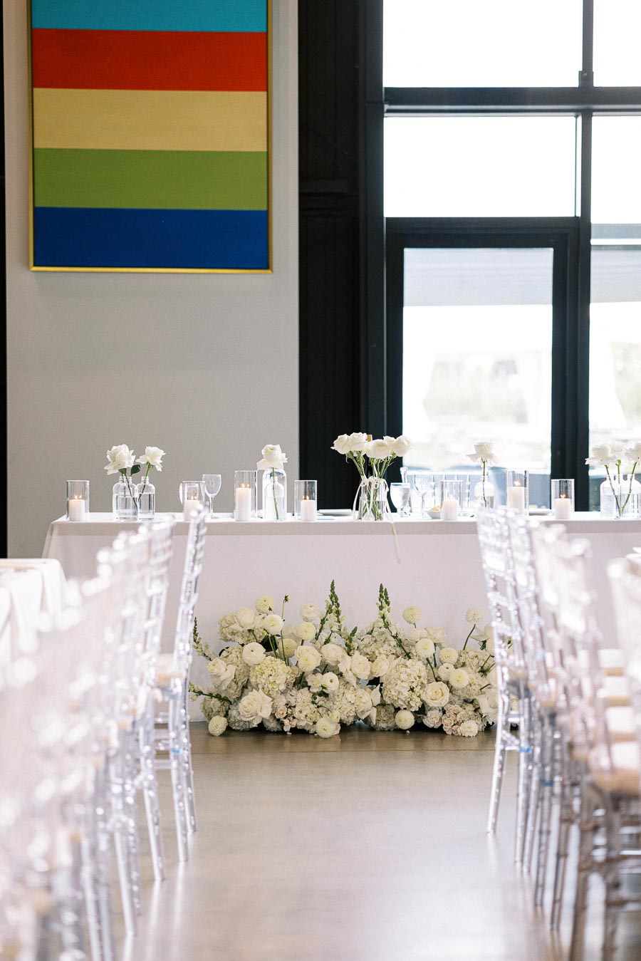 Elegant modern wedding setup featuring white floral arrangements and transparent chairs, with a colorful abstract painting in the background, creating a sophisticated ceremony ambiance.