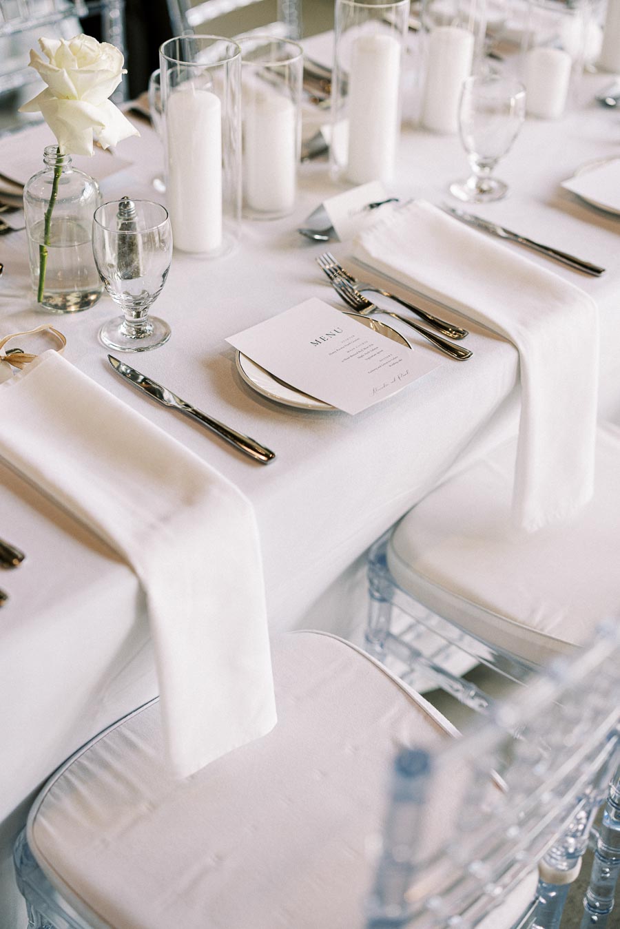 Elegant wedding table setting featuring white linens, glassware, silver cutlery, menus, pillar candles in clear holders, and a single white rose in a vase, creating a sophisticated and romantic ambiance.