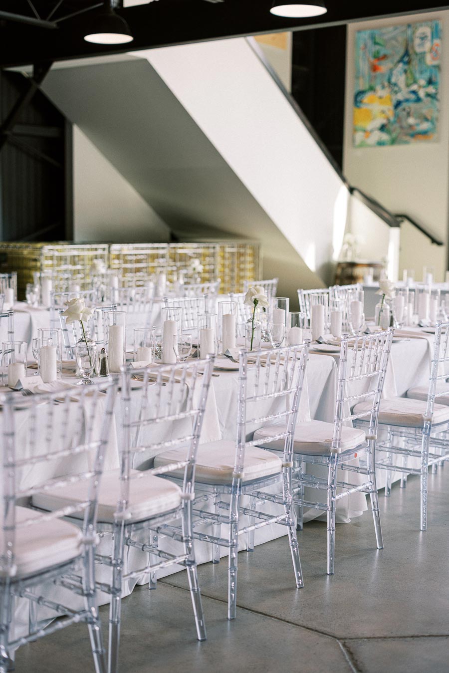 Elegant modern banquet setup in a contemporary event space with transparent chairs, white linens, and neatly arranged table settings featuring white candles and flowers.