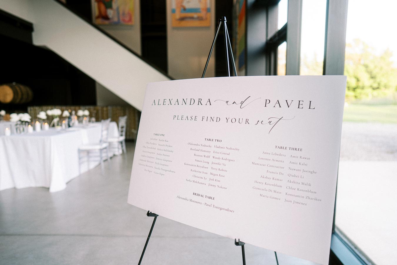 Wedding seating chart on easel for Alexandra and Pavel’s reception, displayed in a modern venue with natural light and elegant table settings in the background.