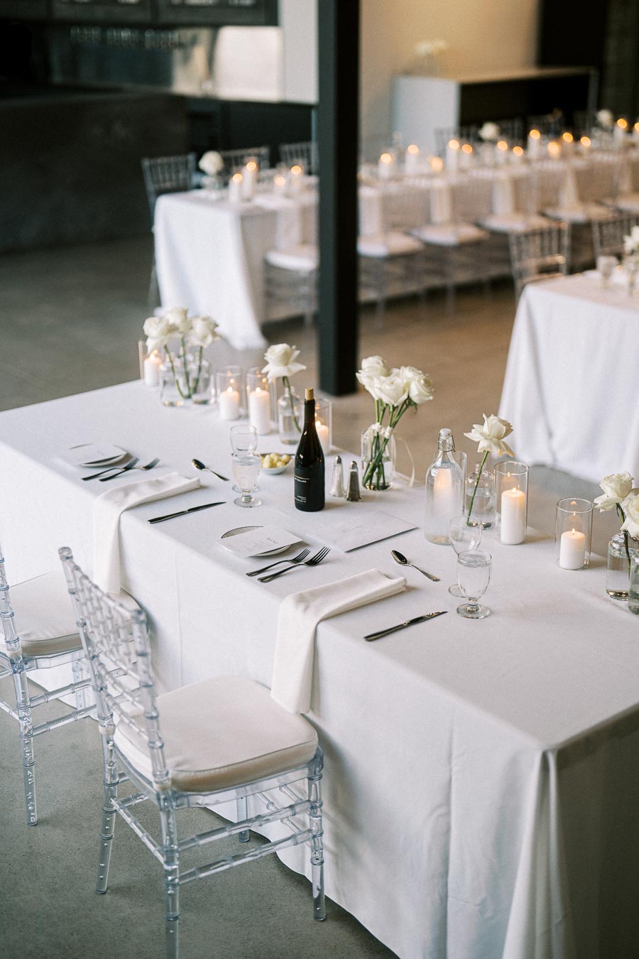 Elegant wedding reception table setting with white tablecloths, clear glass chairs, and candlelit ambiance. Decor includes white roses, wine bottle, and neatly arranged cutlery, creating a sophisticated atmosphere.