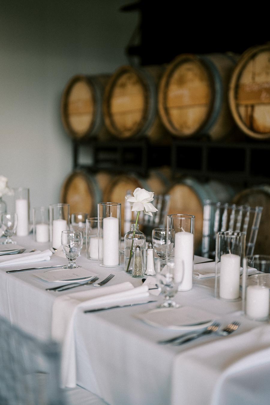Elegant table setting with white tablecloth, candles, and a single white rose, set against a backdrop of wine barrels.