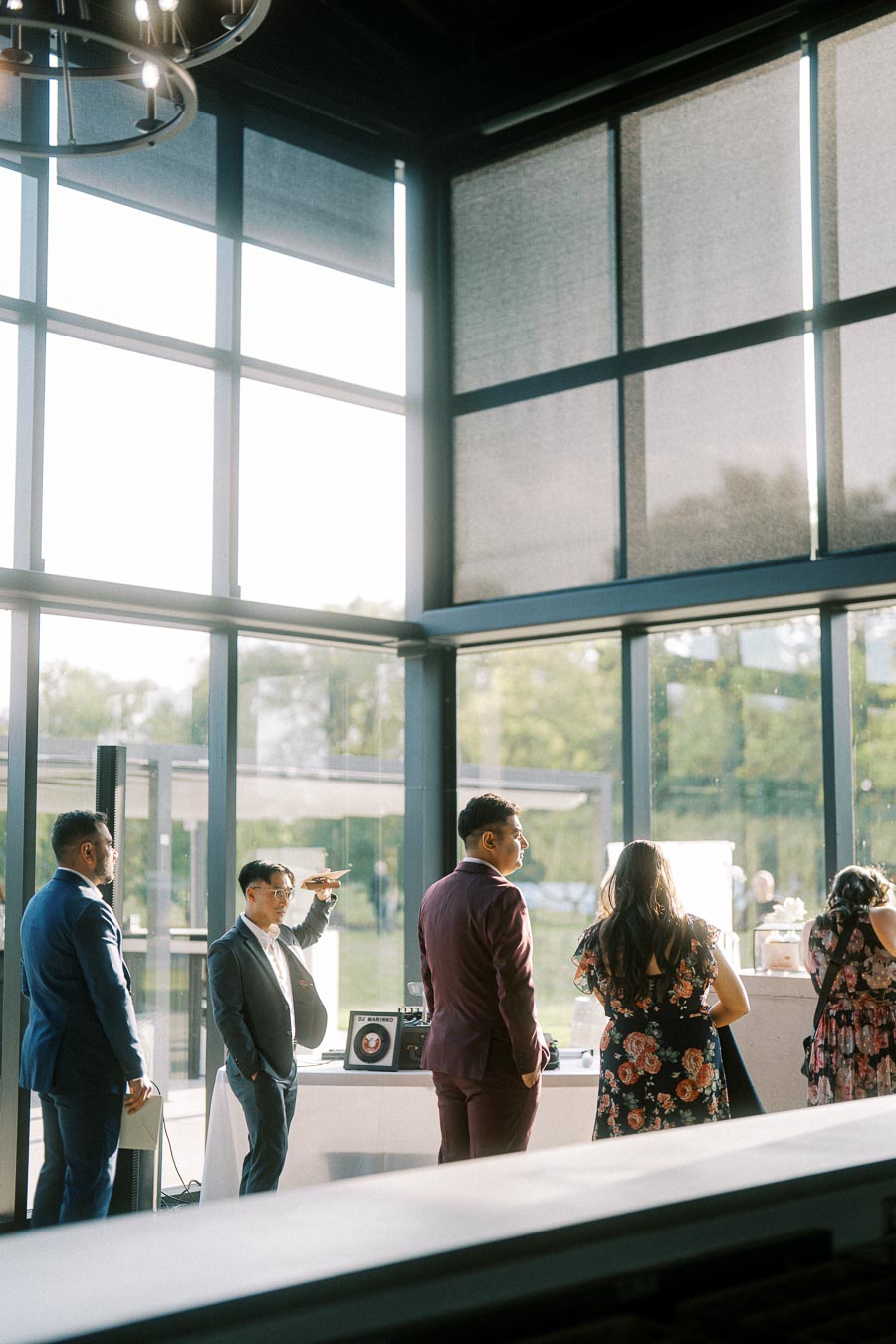 A group of people socializing in a modern, sunlit venue with large windows and a floral arrangement centerpiece reflecting a casual gathering or event atmosphere.