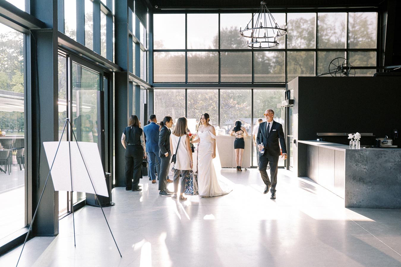 A wedding party gathering in a modern venue with floor-to-ceiling windows, featuring guests mingling and a bride in a white dress, under natural sunlight.