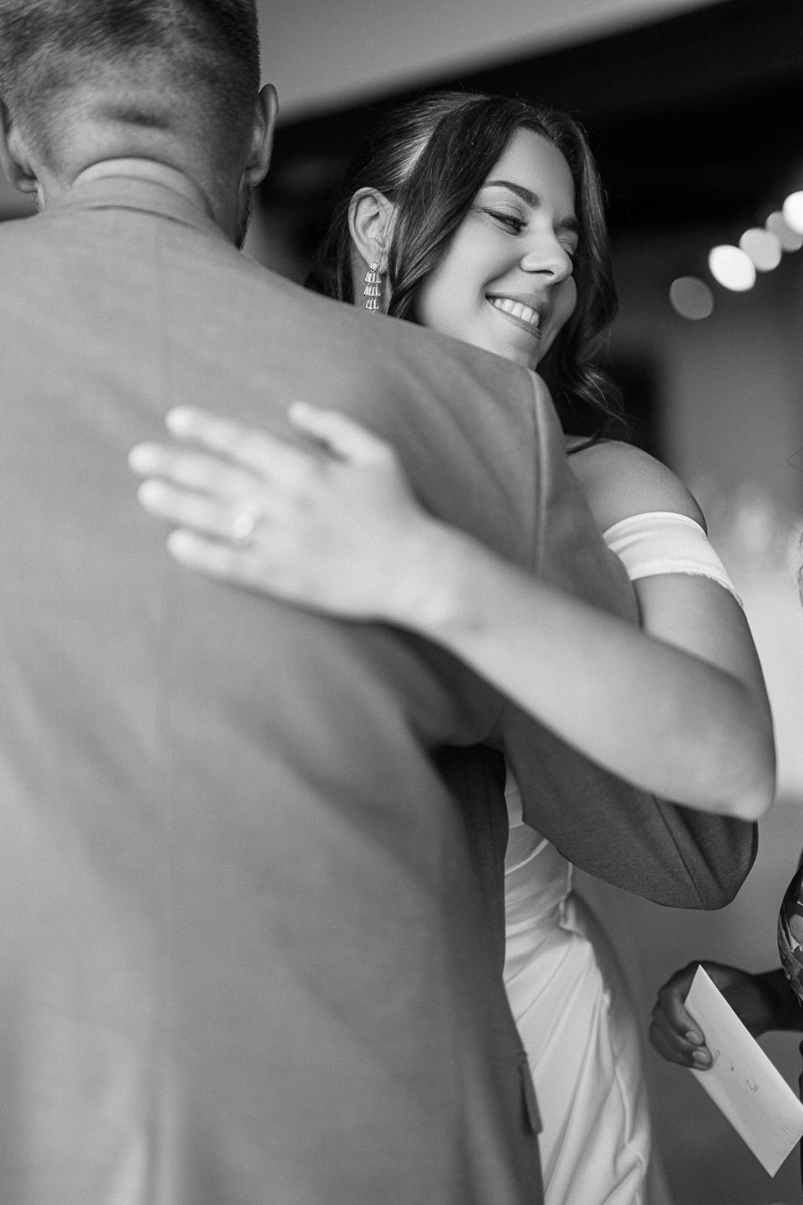 A black and white image of a smiling woman embracing a man, with a close-up on her face. Her expression suggests happiness and warmth, and she is wearing elegant jewelry and an off-shoulder outfit. The scene conveys an intimate and joyful moment.