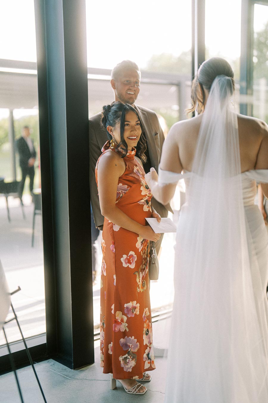 A couple chatting with a bride at a wedding reception, featuring a woman in a floral red dress and a man in a suit, under natural sunlight through large windows.