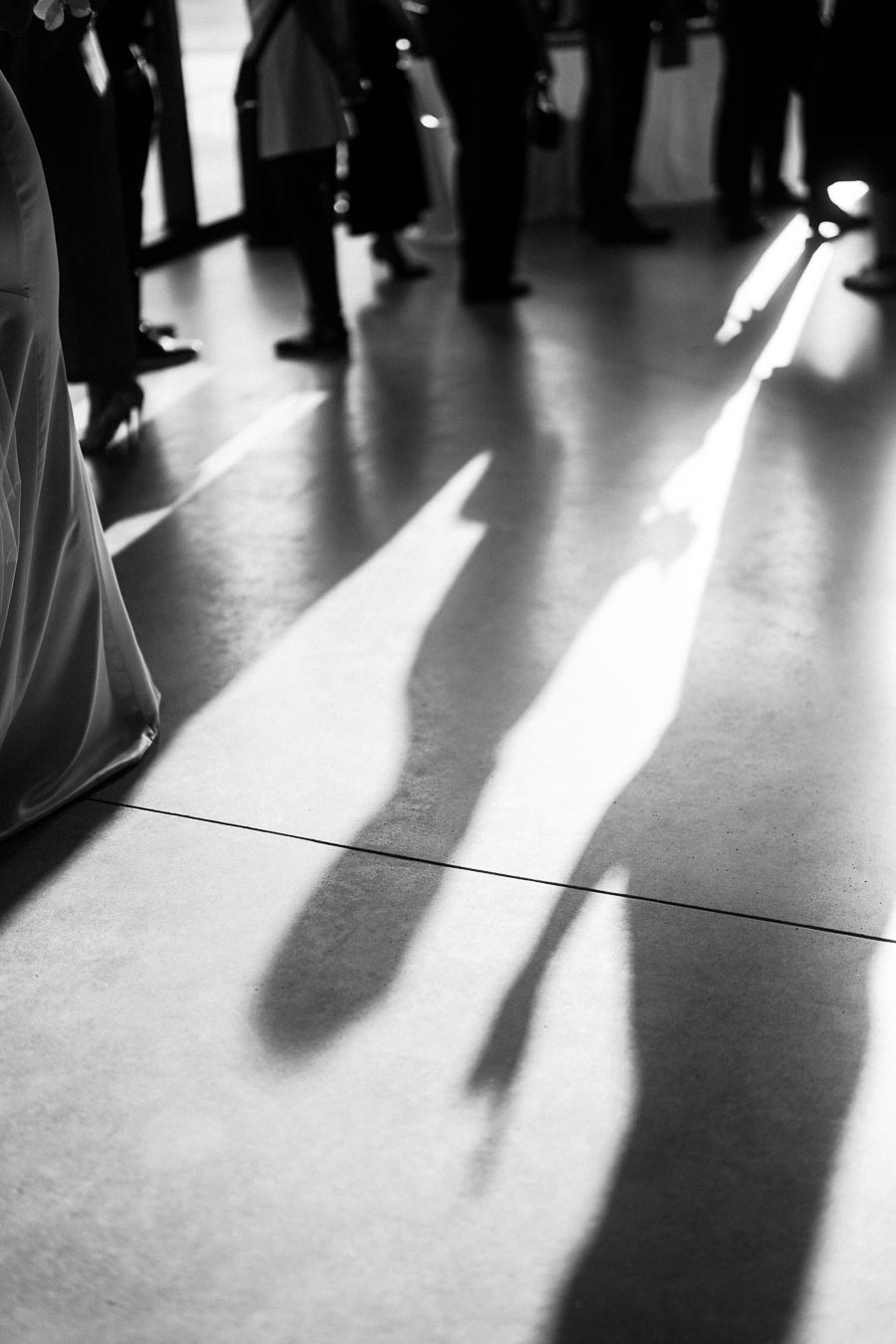 Black and white image showing long shadows of people standing indoors, creating a dramatic contrast on a smooth floor, evoking a sense of mystery and intrigue.