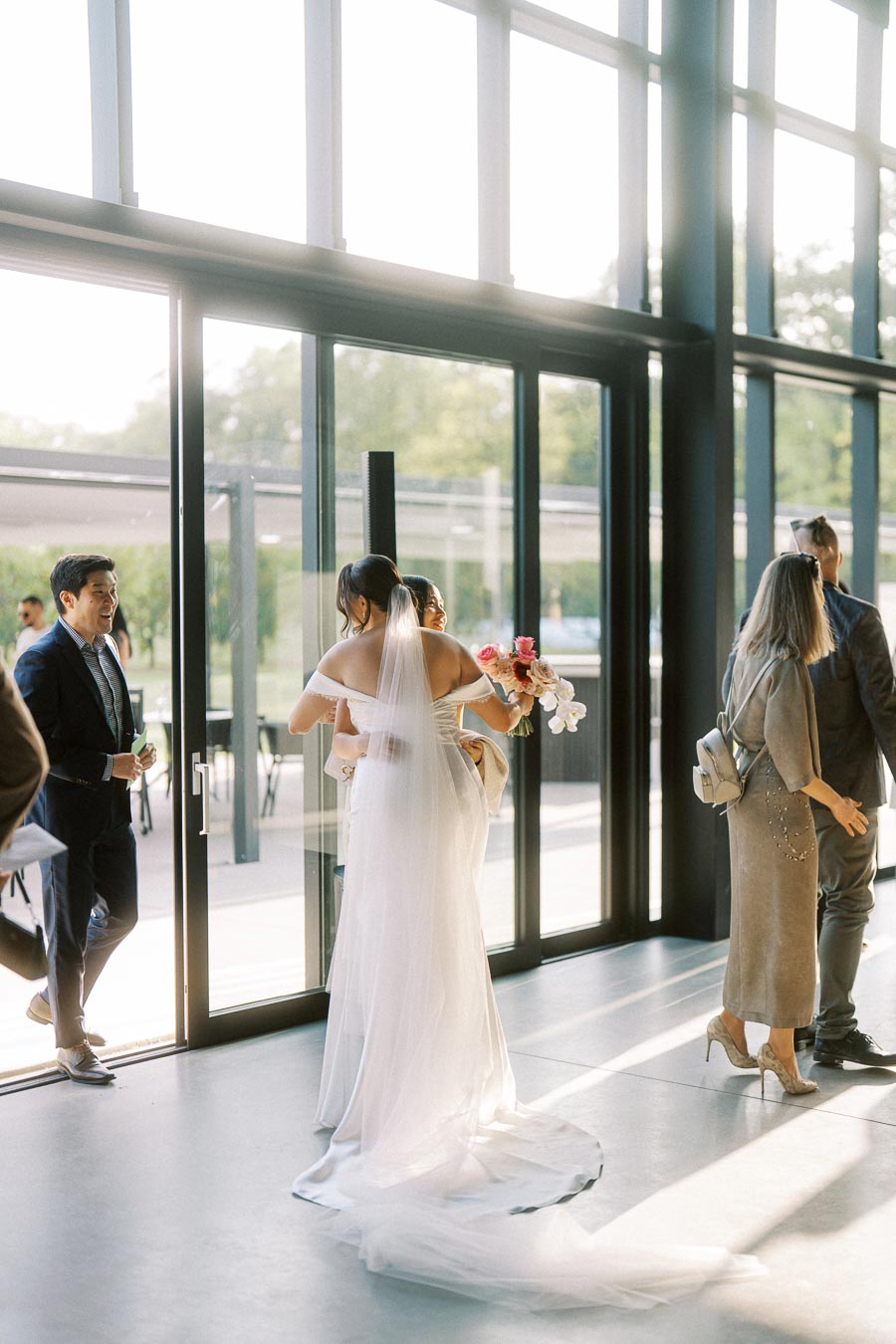 A bride in a long white gown embraces a guest holding a bouquet of pink and white flowers in a sunlit modern venue with large windows, while other guests stand nearby in elegant attire.
