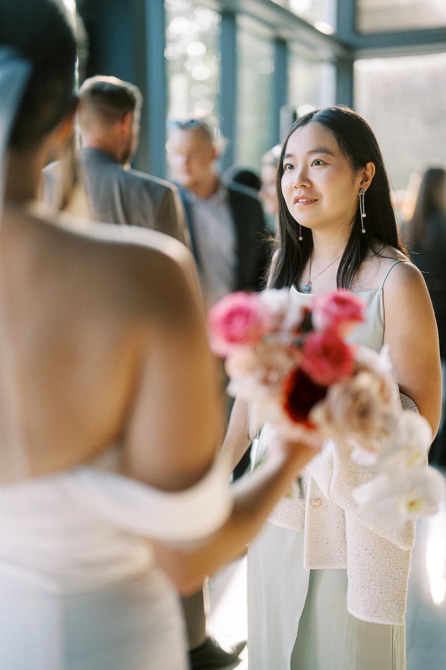 A woman in a light dress smiles while talking to a bride holding a bouquet of flowers, in a sunlit indoor setting with blurred guests in the background.