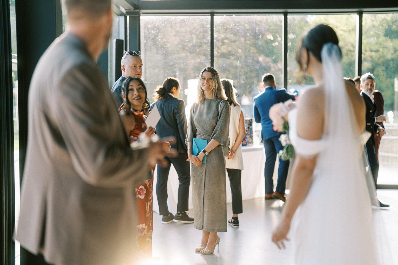 A diverse group of wedding guests socializing in a sunlit venue, with a woman in a gray dress holding a bright blue clutch and a bride in white slightly out of focus in the foreground.