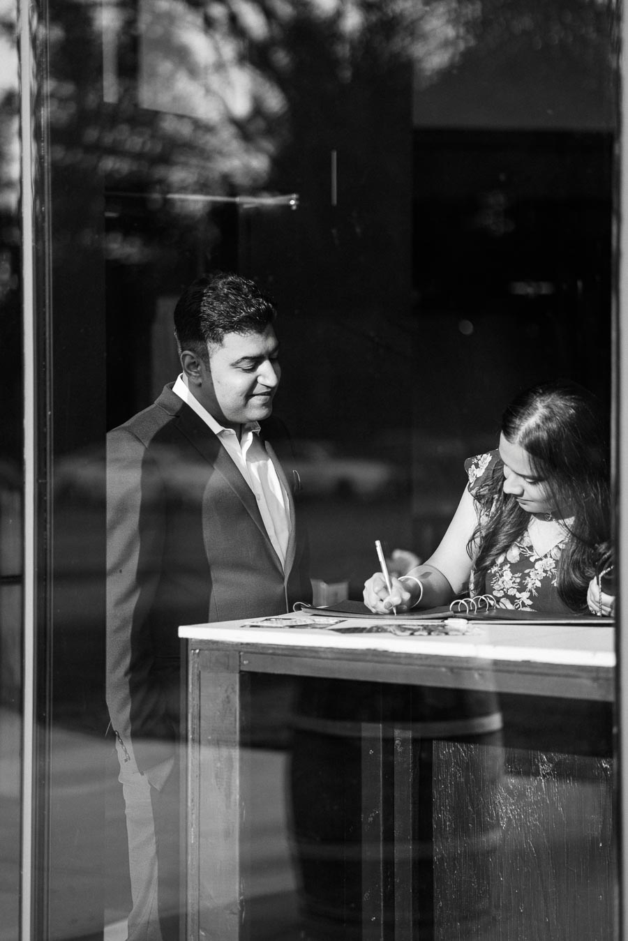 Black and white photo of a man in a suit standing next to a woman writing in a notebook, viewed through a glass window with reflections and shadows.