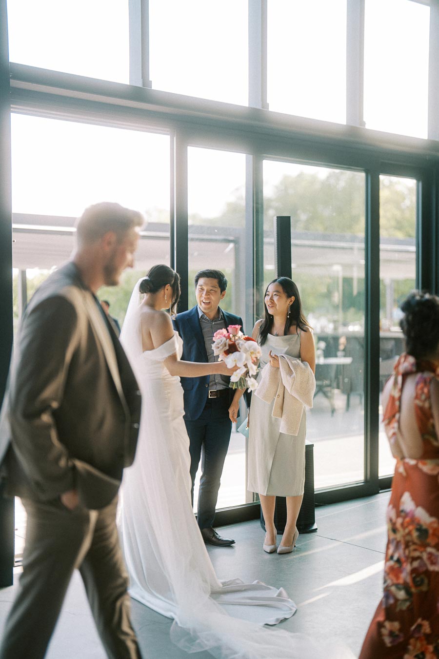 Bride in an elegant white gown with a long veil holding a bouquet and chatting with guests at a modern wedding venue with large windows.