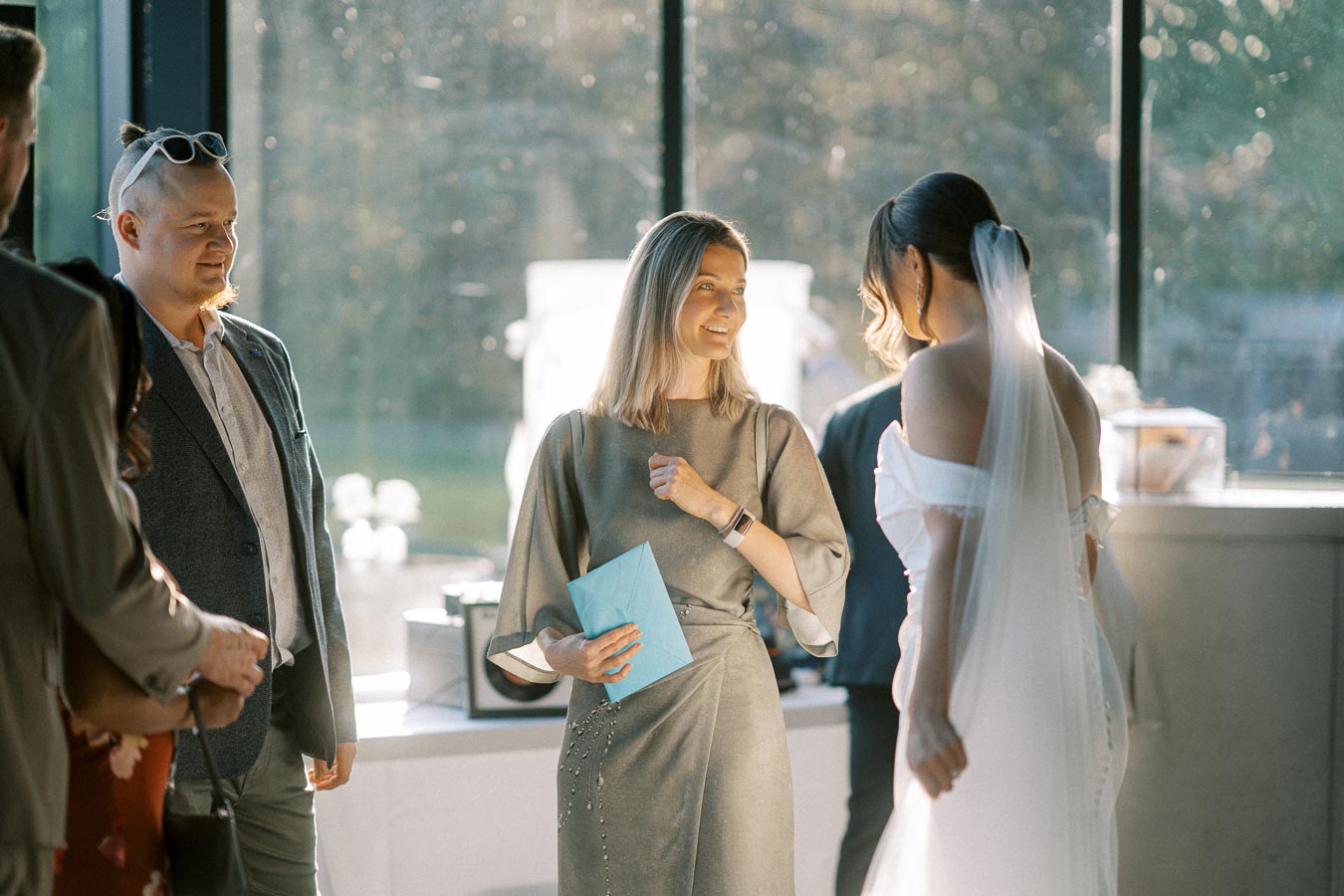 Group of people at a wedding, with one person holding a blue envelope and another wearing a white wedding dress with a long veil, standing in front of large windows.