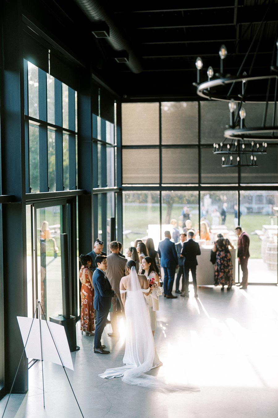 Elegant wedding reception in a modern venue with large windows, guests mingling in stylish attire, and a bride in a flowing white gown.