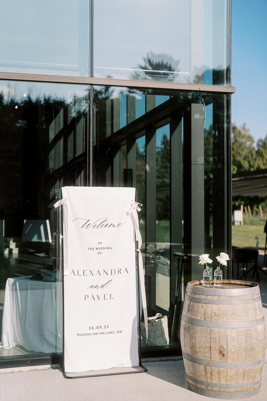 Outdoor wedding venue entrance with a welcome sign for a wedding ceremony featuring a couple's names, Alexandra and Pavel. The sign is propped on a stylish stand outside a modern glass building, with a rustic wooden barrel and flowers nearby.