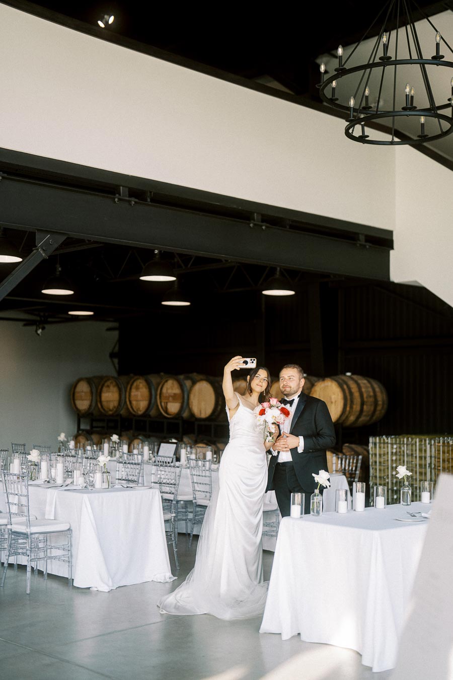 Couple taking a selfie at a wedding venue with wine barrels and elegant table settings in the background.