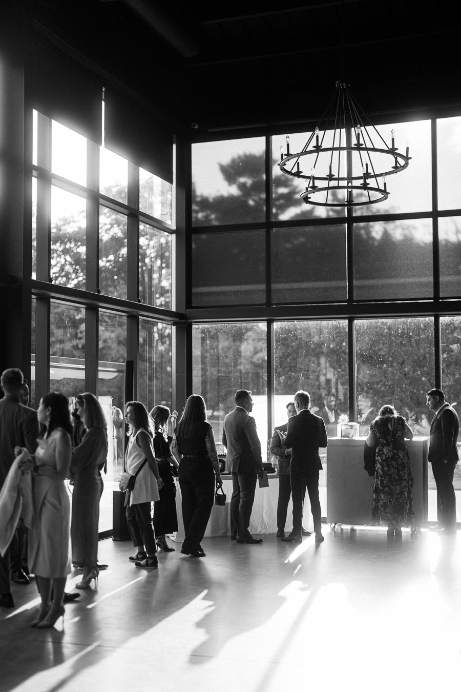 Black and white image of a social gathering in a modern venue with large windows and a chandelier, showing people in formal attire engaged in conversation and networking as sunlight streams through the windows.